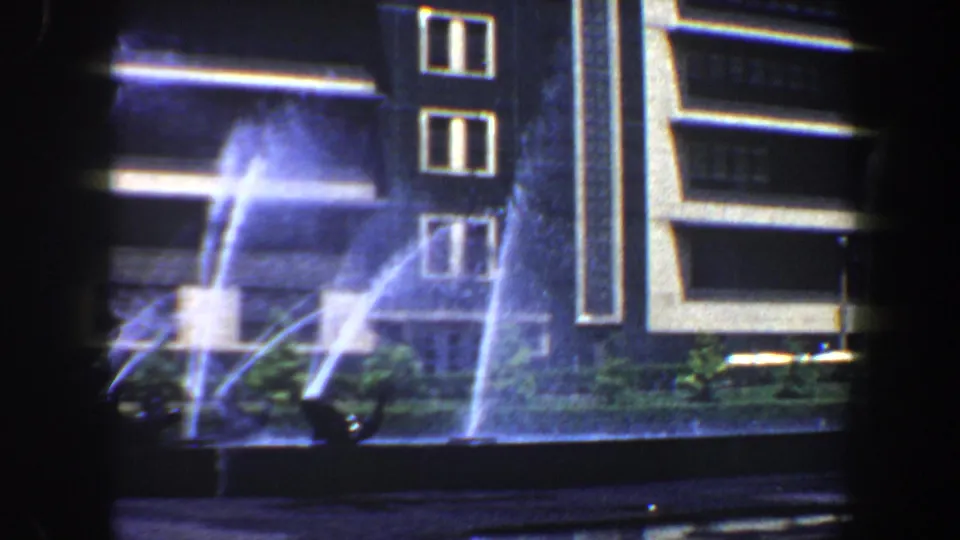 1964 Saint Louis frame of fountain jets crossing a landscaped basin beside a modern building and street traffic.