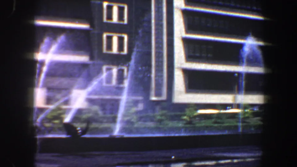 1964 Saint Louis home-movie frame showing a illuminated building exterior, fountain spray, planter, and dark foreground framing.