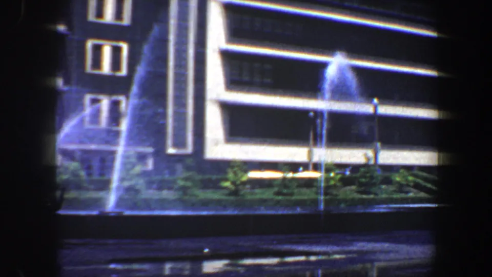 Nighttime 8mm view in Saint Louis with a modern building facade, blue fountain spray, street edge, and landscaped planter.