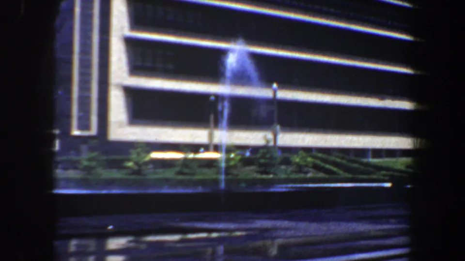 1964 Saint Louis frame with fountain water, illuminated architectural lines, greenery, and wet pavement from a handheld 8mm view.