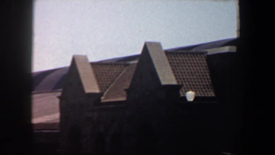 1964 Saint Louis 8mm frame showing tiled gable peaks and a grey rooftop with soft grain and heavy foreground shadow.