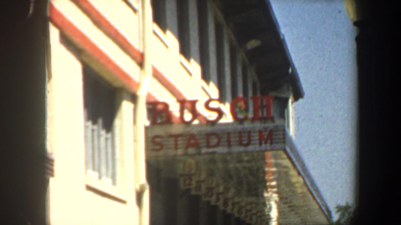 Sunlit building facade with red BUSCH STADIUM lettering and blue sky at right.