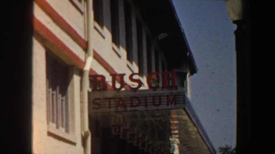 1964 Saint Louis home-movie frame with the BUSCH STADIUM sign framed beside pale wall surfaces, windows, red trim, and dark edge shadows.