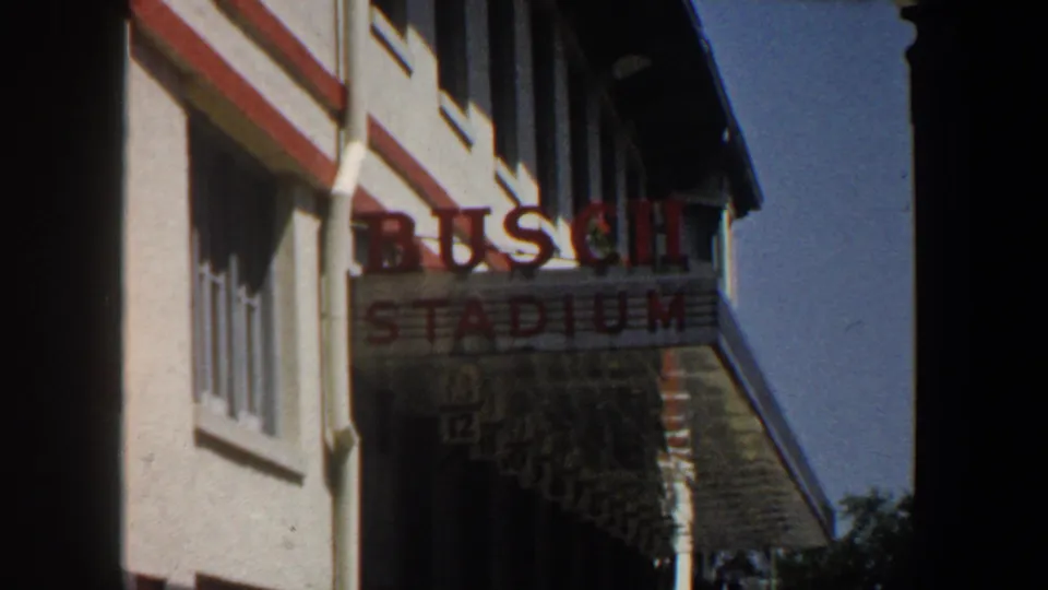 1964 Saint Louis frame holding on the BUSCH STADIUM sign, with the building facade, windows, angled roofline, blue sky, and dark frame edge visible.