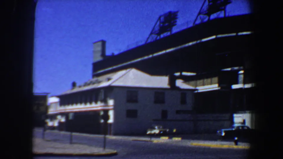 1964 Saint Louis roadway beside a white building and elevated stadium structure, with cars moving or parked along the curb.