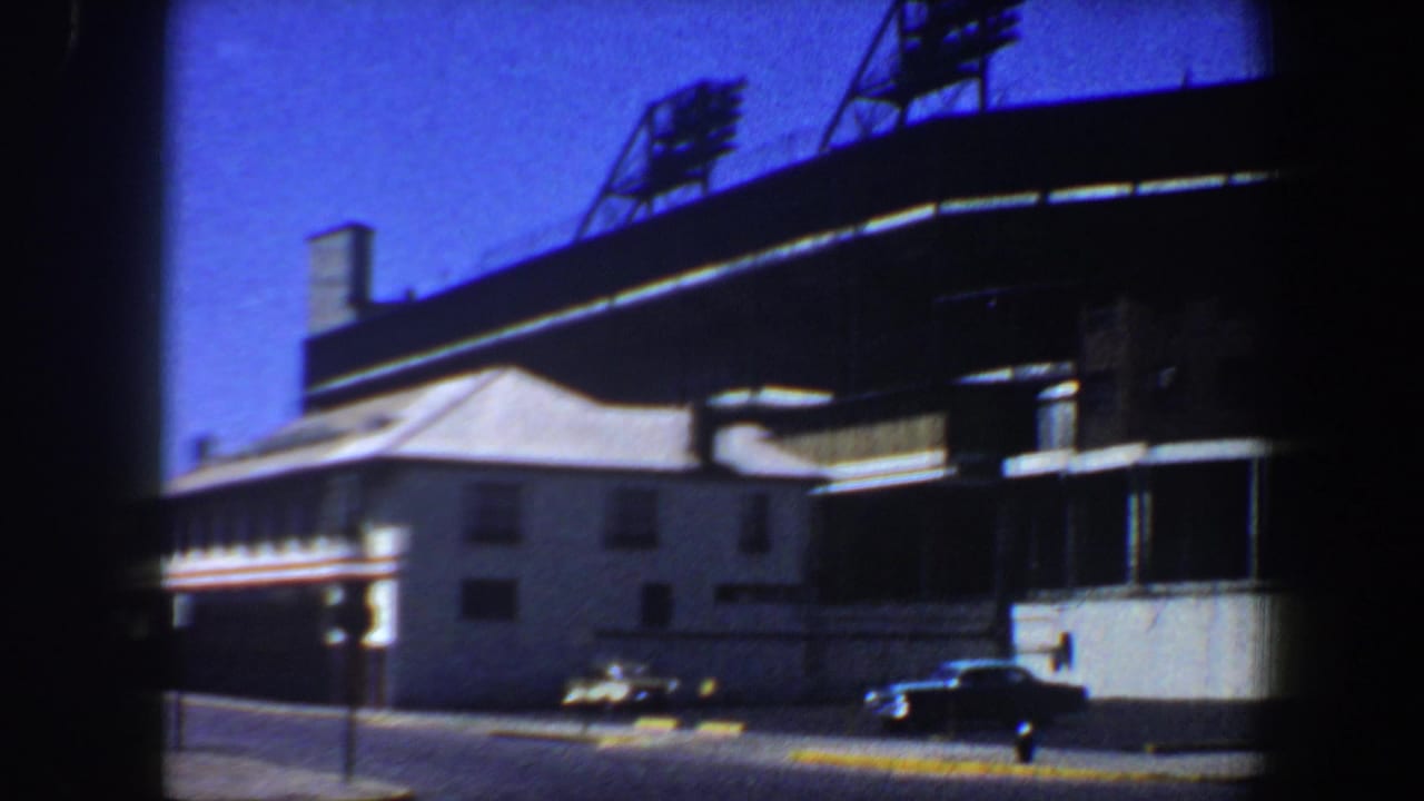1964 Saint Louis 8mm frame showing the exterior of a Cardinals baseball stadium, with light towers above the roofline and cars along the street.