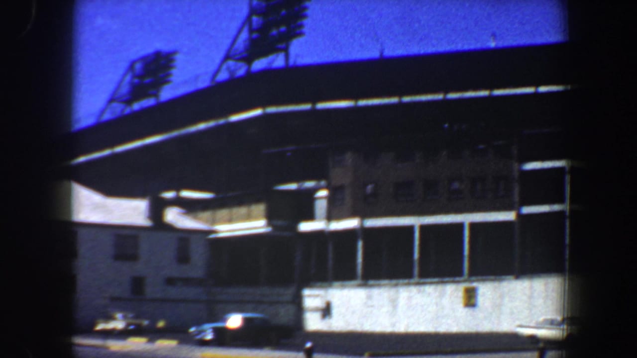 Closer stadium exterior view with a signboard area, light towers, and cars near the curb.