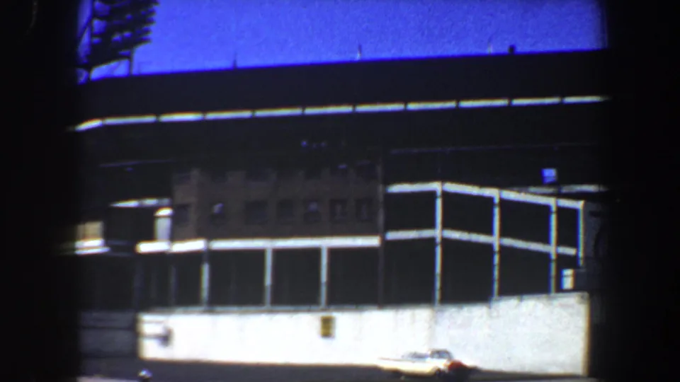1964 Saint Louis 8mm frame of the stadium facade and corner, with white lower wall panels, dark upper structure, and a parked car near the building.