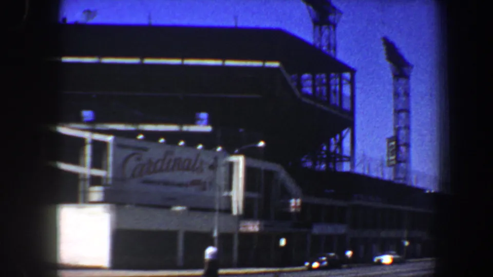 A moving-vehicle view in St. Louis shows Cardinals signage on a large complex with period cars along the street.