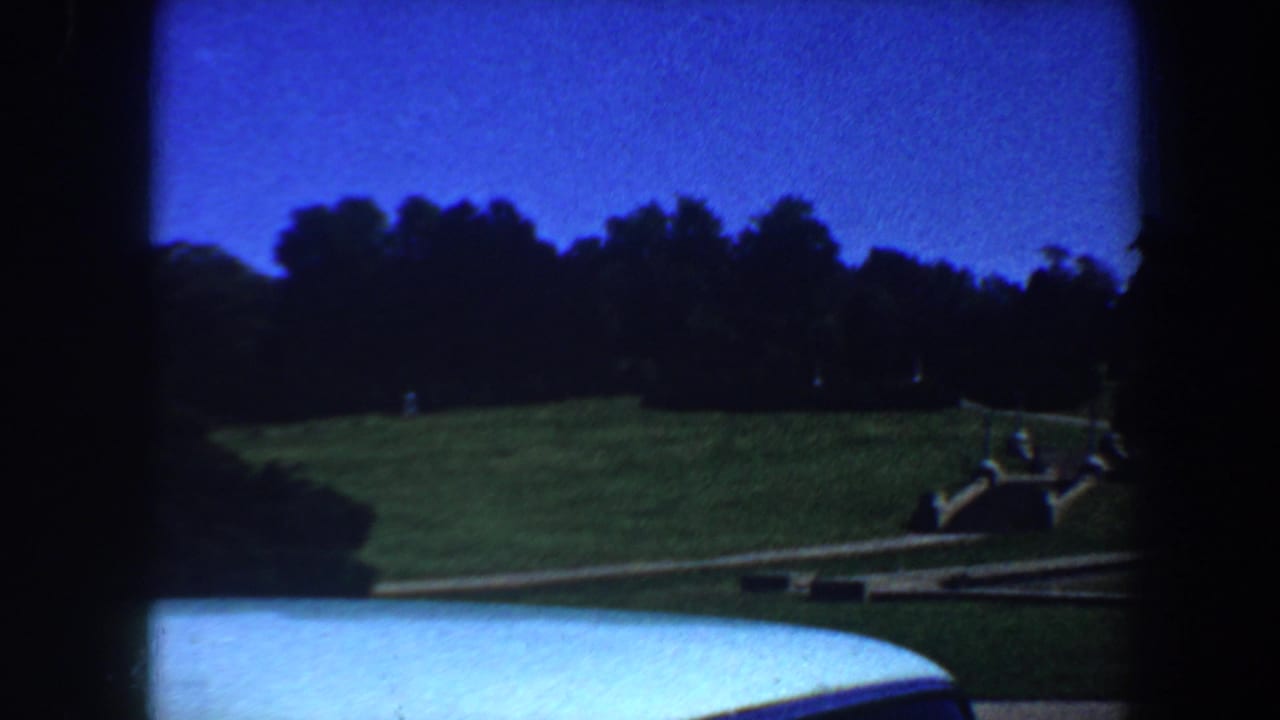 Car-window-framed view of a lawn, water edge, walkway, trees, and distant figures.