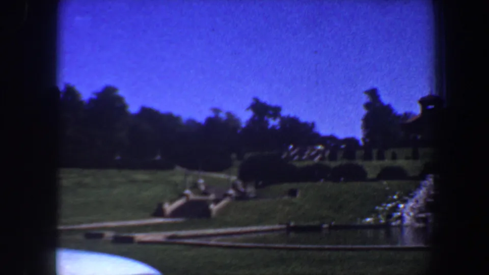 1964 Saint Louis frame showing a wider landscaped view with water in front, a path across the lawn, shrubs, trees, and distant visitors.