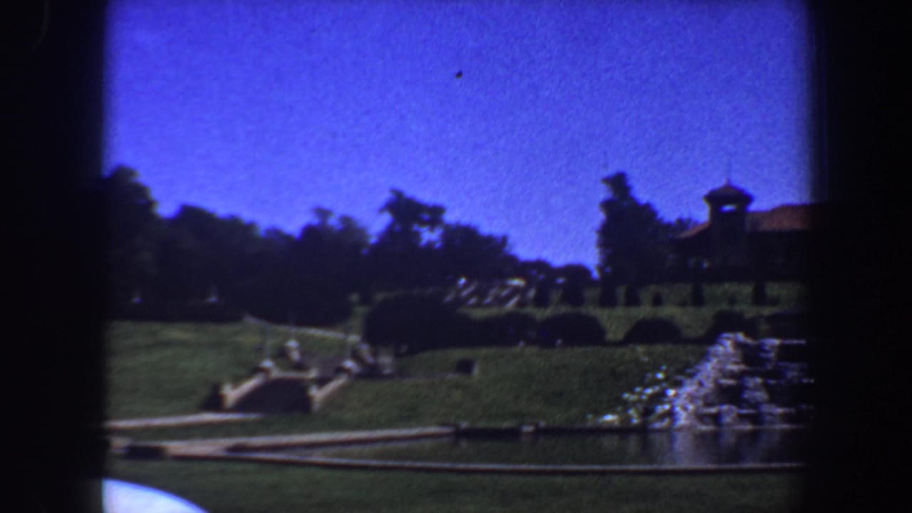Formal brown building beyond green lawns, flower beds, and a cascading fountain under a blue sky.