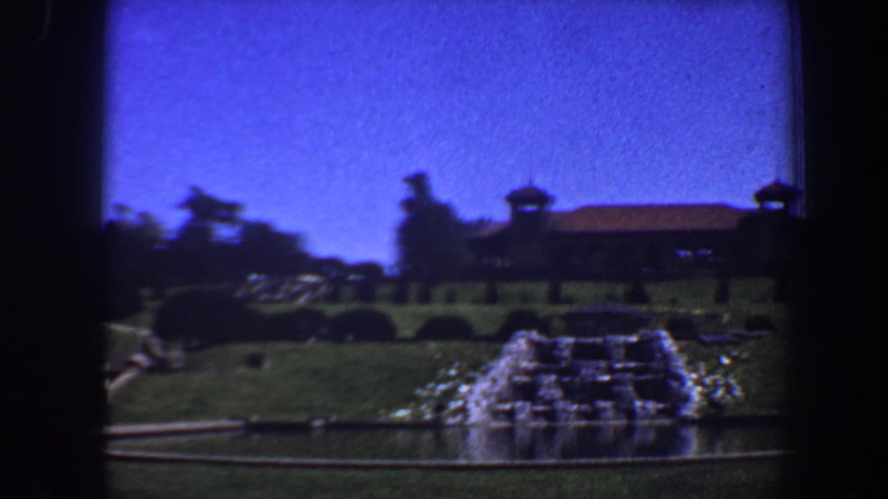 Cascading fountain and pool in front of a distant brown building with turret-like roof forms.