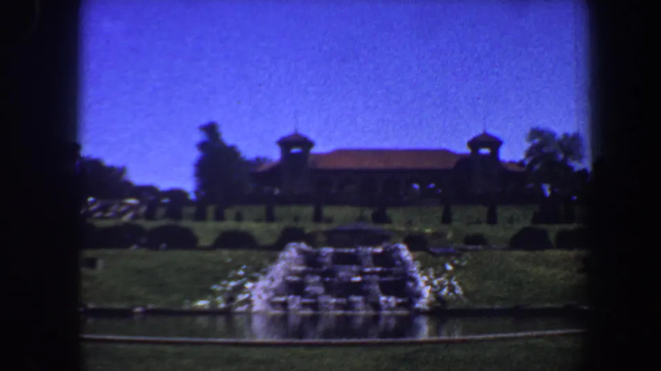 Home-movie frame from 1964 Saint Louis with a centered fountain, formal lawn, and distant brown building with twin turrets.