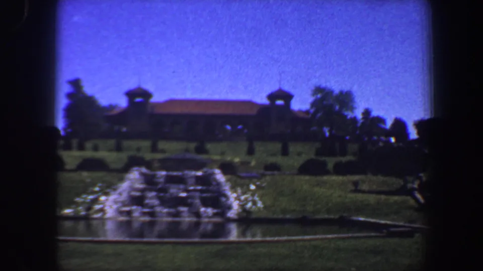 1964 8mm Saint Louis frame showing a soft-focus fountain scene with manicured greenery and a distant brown building.