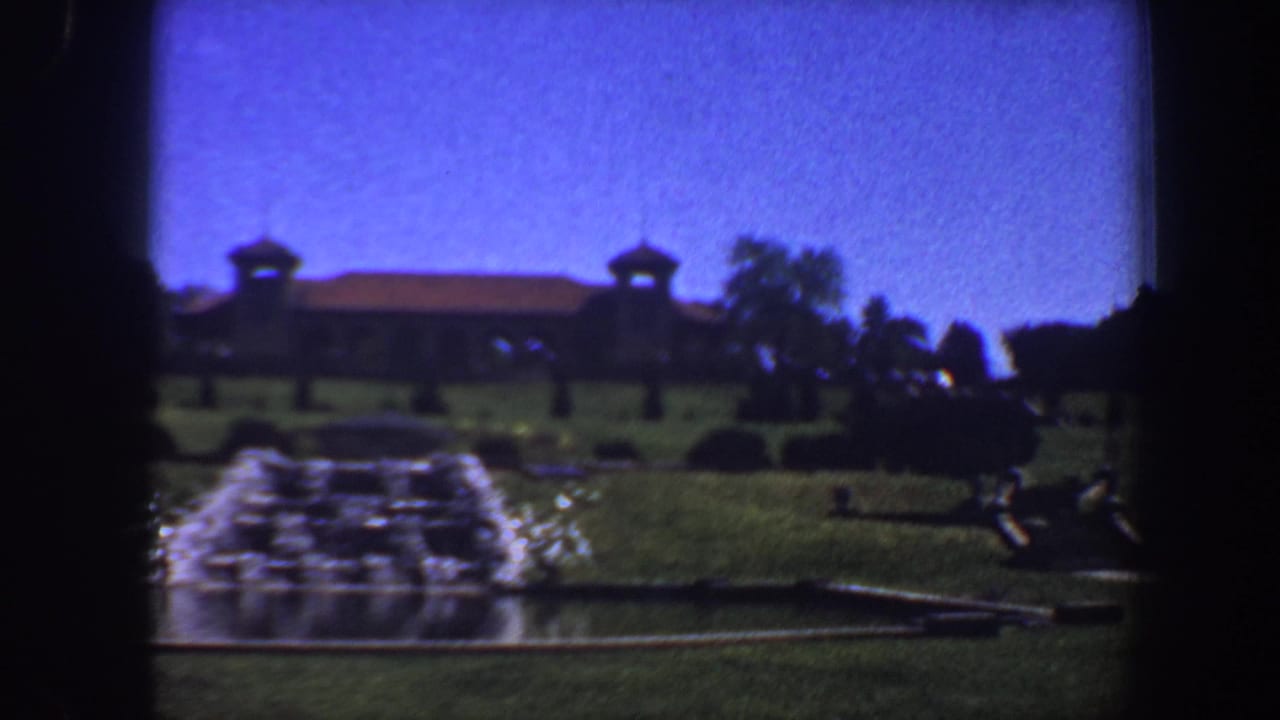 Manicured lawn with fountain spray, distant building, trees, and blue sky.