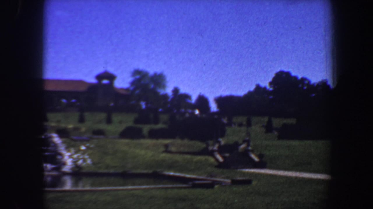Green lawn, fountain edge, path, trees, and distant building in sunlight.