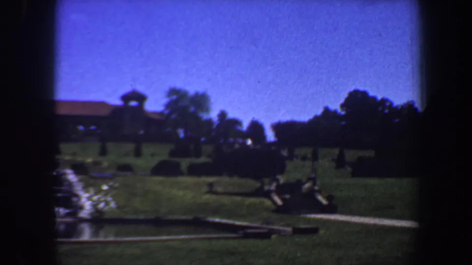 1964 Saint Louis 8mm frame with the camera shifted right across lawn, fountain water, a path, trimmed shrubs, trees, and the distant building.