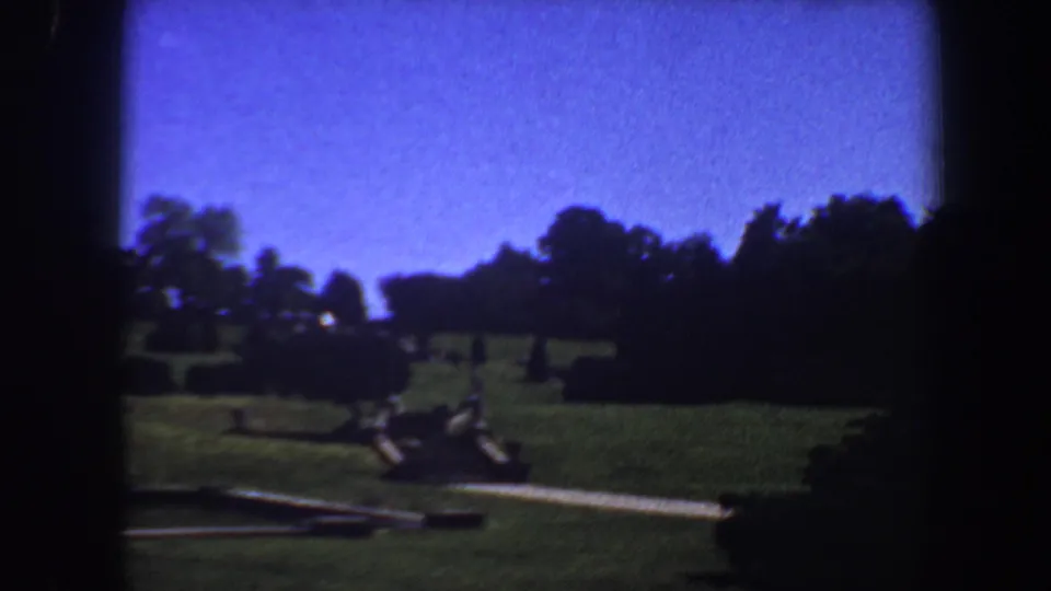 1964 Saint Louis 8mm frame showing a darker sweep of lawn, curving path, trimmed shrubs, tree line, and blue sky as the building falls mostly out of frame.