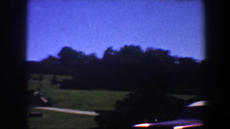 1964 Saint Louis 8mm frame with lawn, path, shadowed trees, shrubs, blue sky, and a partial car body entering the lower right edge.