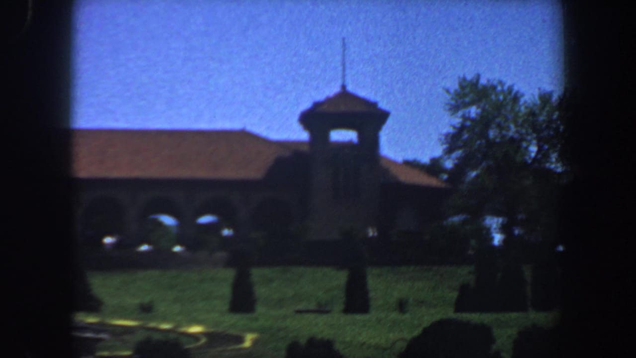 Long arched building and tower seen across a sunlit lawn with trees at right.