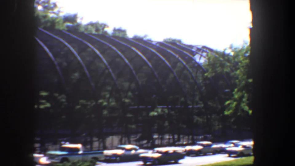A side angle shows the arched park structure behind trees, with multiple period cars parked or passing in the foreground.