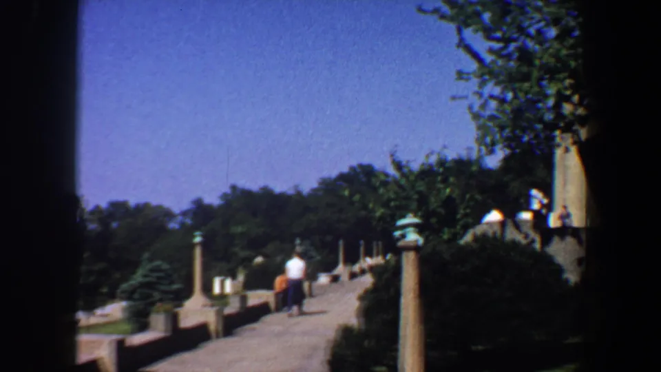 A Saint Louis park walkway shows columns, shrubs, trees, and a few distant visitors under bright daylight.