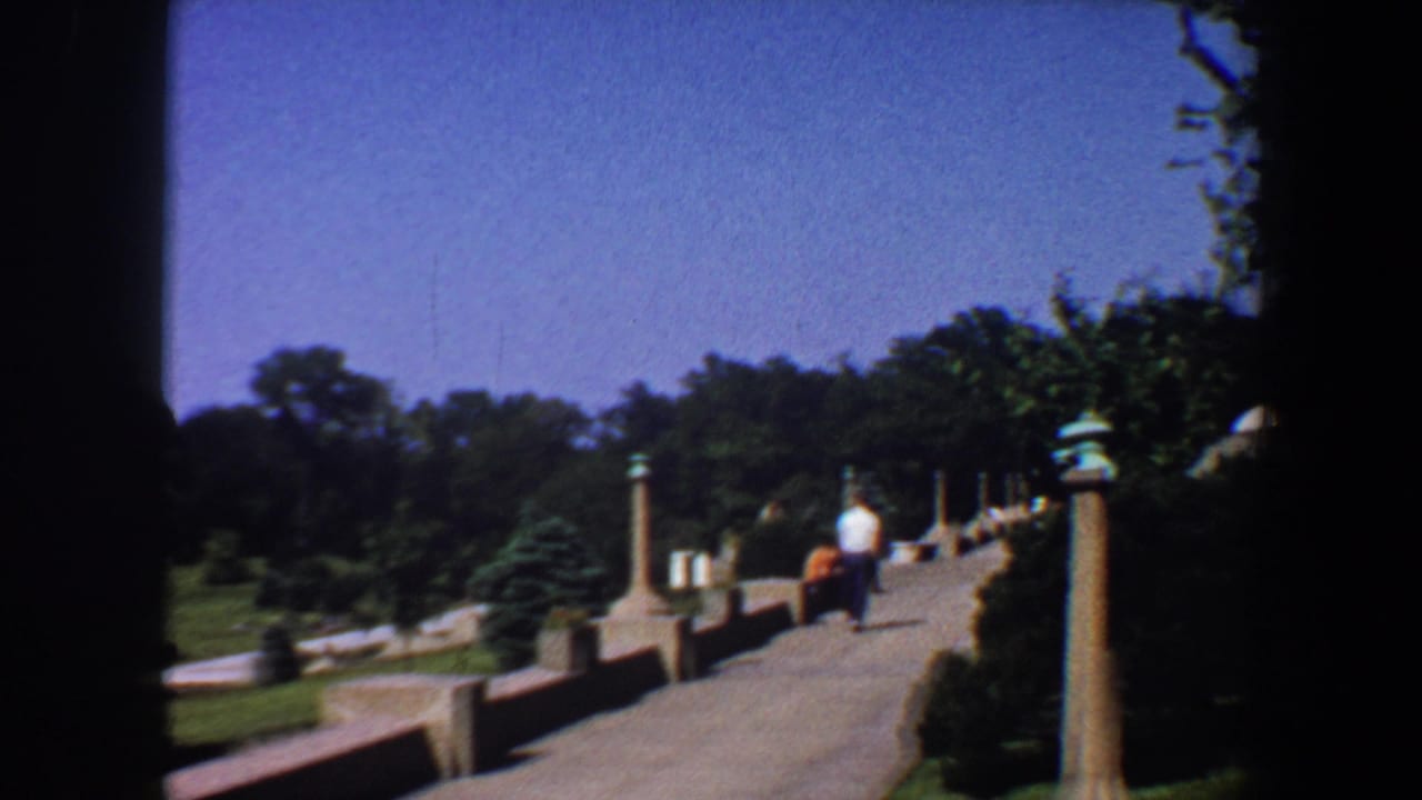 Paved garden path with low walls, ornamental pillars, lawns, trees, and a distant walker.