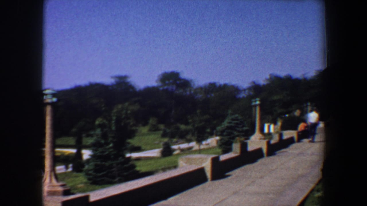 Garden walkway and lawn seen past a dark film edge, with pillars, trees, and a distant person.