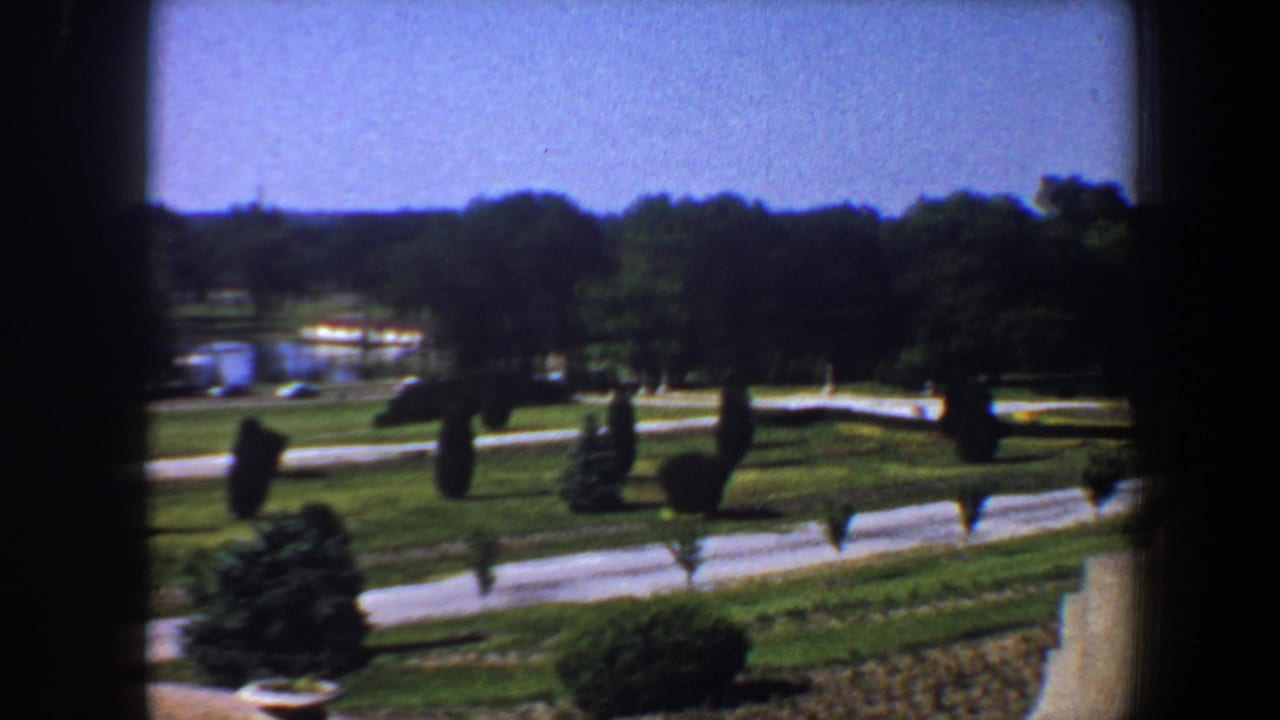 Park grounds seen through a dark frame, with roads, lawns, hedges, trees, and water in the distance.