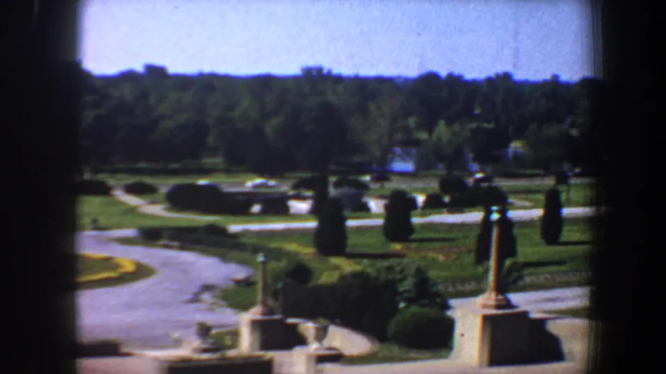 1964 home-movie frame of a Saint Louis park roundabout area, with lawns, paved paths, hedges, stone posts, trees, and distant lake reflections.
