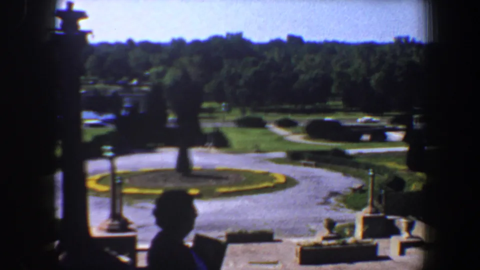 Saint Louis park view from 1964 8mm footage, showing curved roads, lawns, hedges, trees, stonework, and a silhouetted person near the lower right.