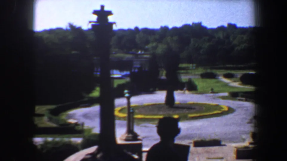 1964 Saint Louis home-movie frame showing a circular drive, manicured garden beds, and a silhouetted visitor near a shaded estate entrance.