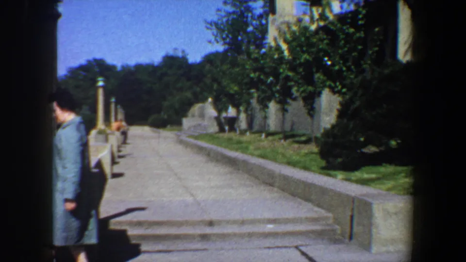 The woman passes the edge of the frame as the camera view opens onto steps, a stone path, lawn, shrubs, and tree-lined garden space.