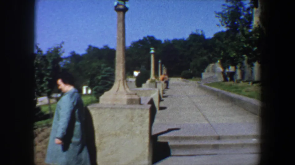 A woman in teal walks by a tall stone post and low wall on a sunlit Saint Louis garden pathway in 1964.