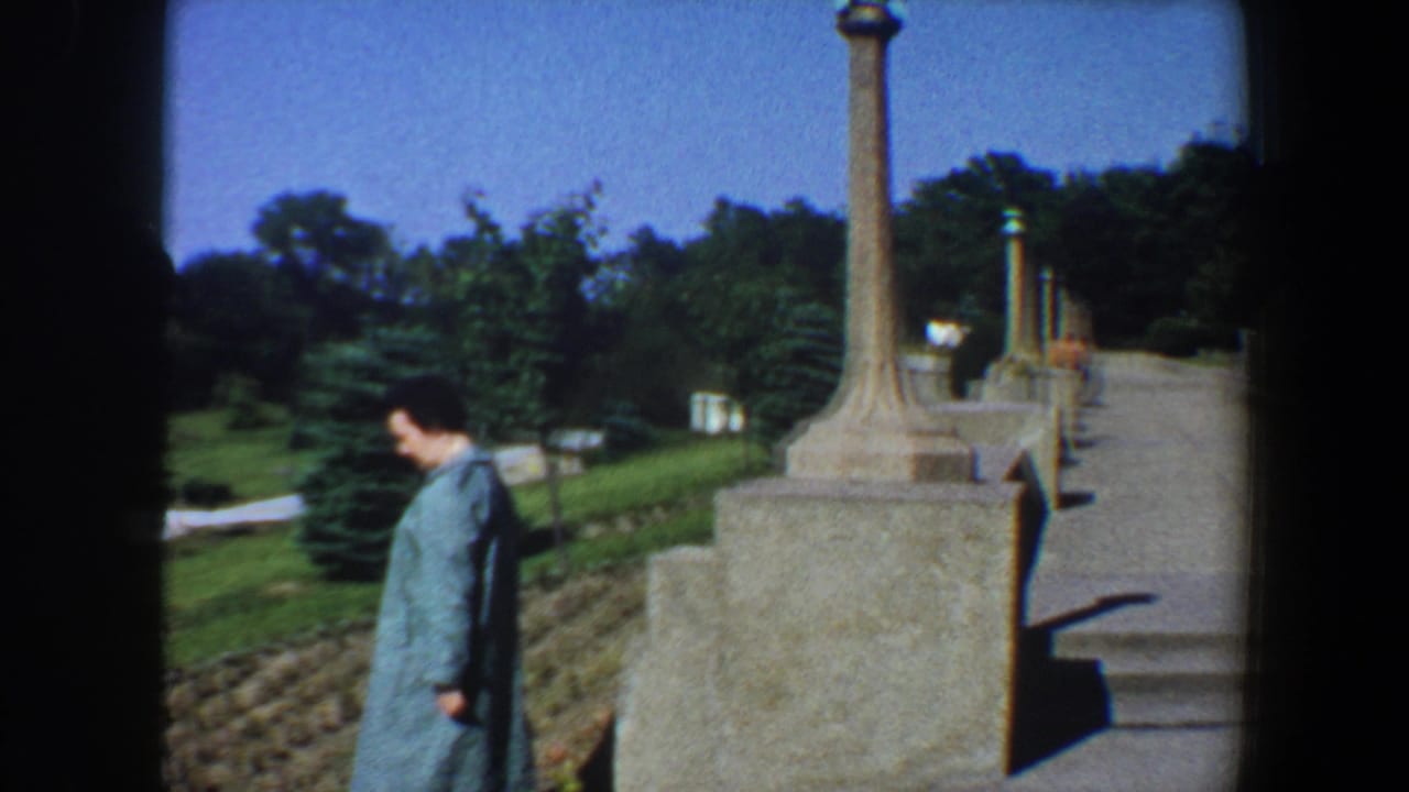 Woman in a blue coat walking beside stone pillars and planted beds in a sunny park.