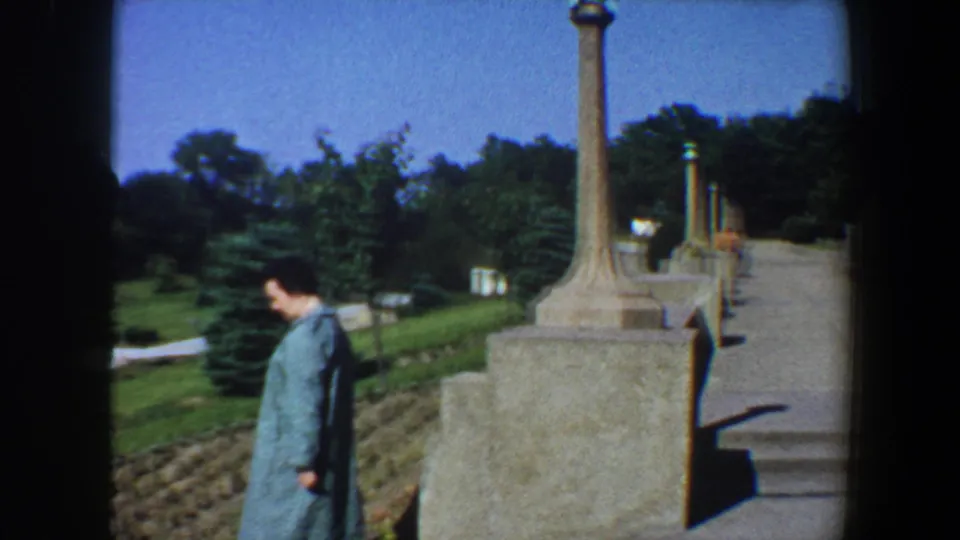 In Saint Louis, Missouri in 1964, a woman in a long blue coat walks near stone pillars, sloped planting beds, lawns, and trees.