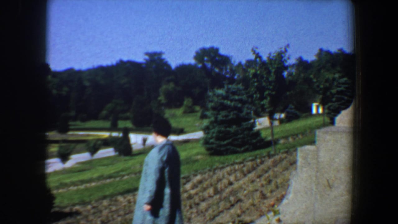 Woman in a blue coat walking downhill near lawns, shrubs, and a stone pillar.