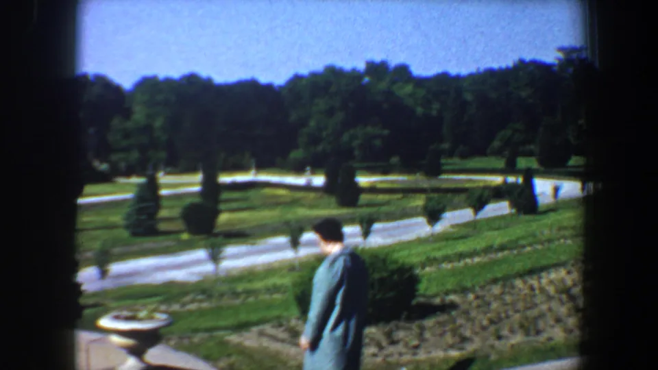 The woman in a blue coat walks along the landscaped Saint Louis grounds, with a stone planter, clipped shrubs, curved paths, and trees visible.