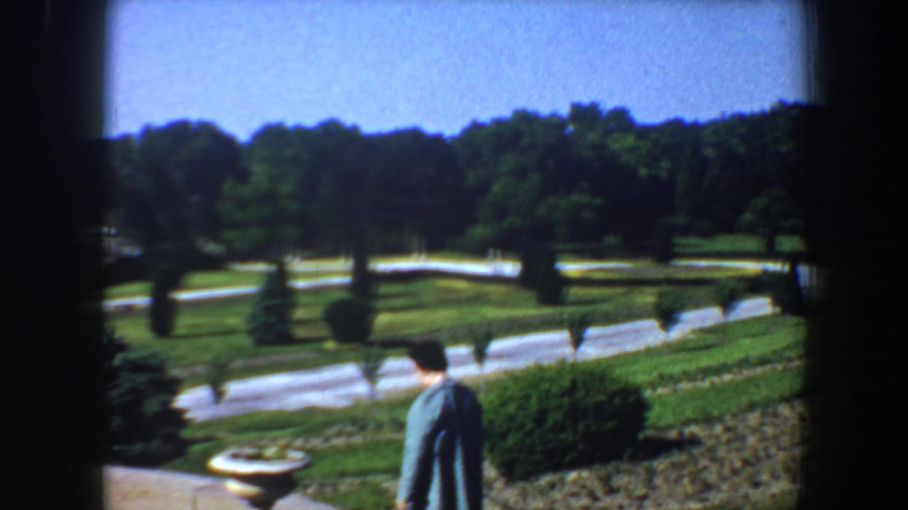 Woman in a blue jacket walking past planters above a formal garden with lawns and paths.