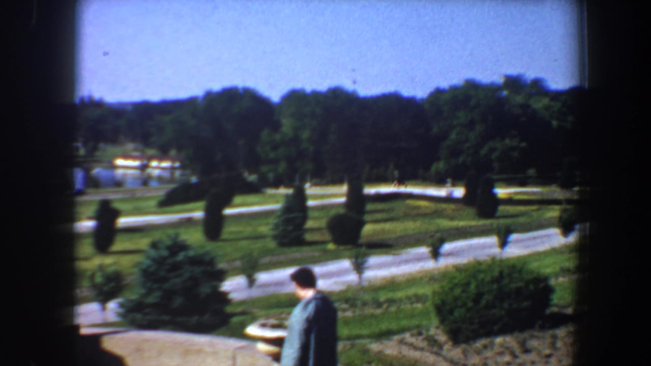 Woman walking along the lower edge of a landscaped garden with winding paths and trees.