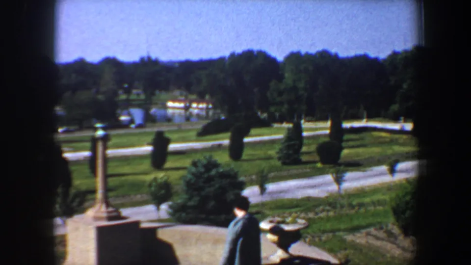 The 1964 Saint Louis home-movie frame shows a woman near a planter, with paths, lawns, trees, and a column-like garden feature.