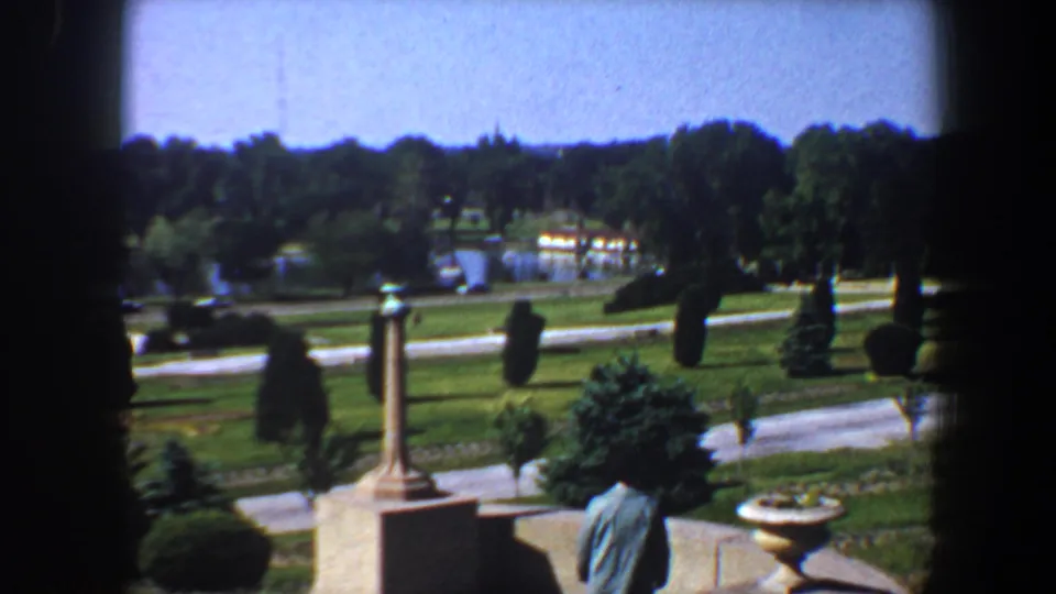 A woman in a blue jacket is seen from behind in supplied 1964 Saint Louis footage, with formal paths, shrubs, trees, and distant water.