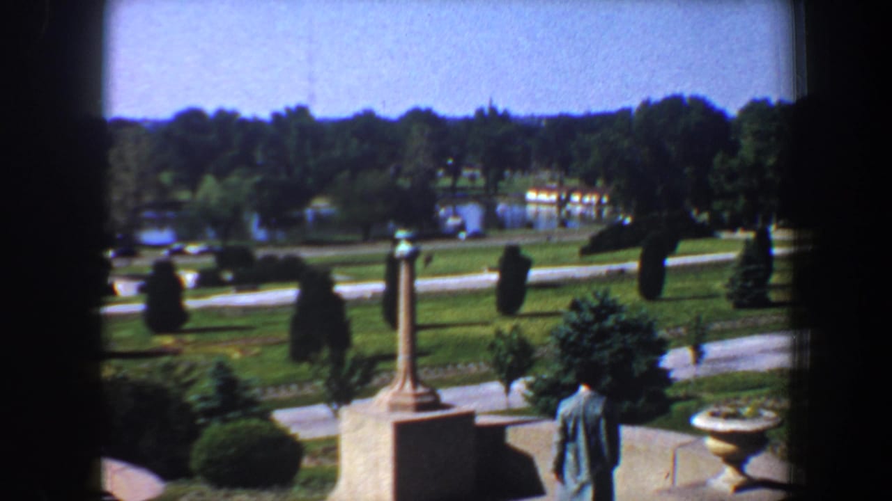 Wide park view with lawns, paths, trees, distant water, and a columned statue.