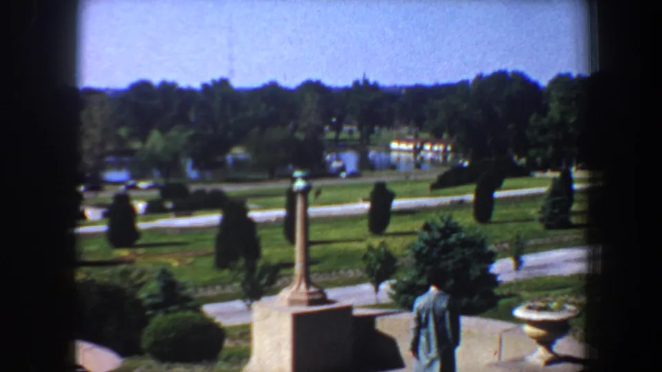1964 Saint Louis park view with manicured lawns, curving paths, trees, distant water, and a columned monument in the foreground.