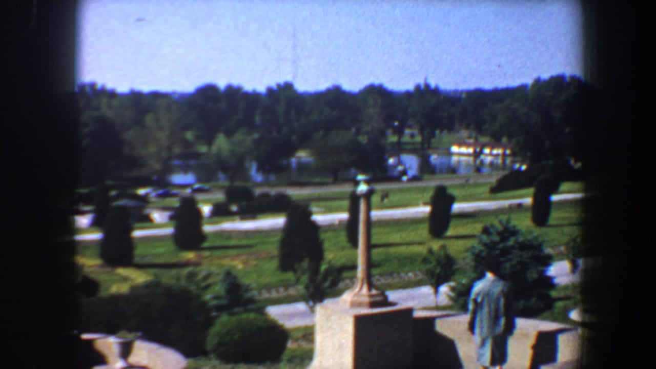 Man in a coat and hat walks near a statue overlooking a park landscape.