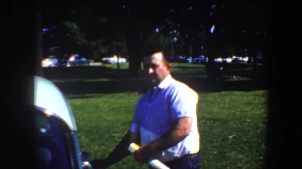 Adult man in a light short-sleeve shirt stands beside a dark car on grass, with trees and parked cars behind him.