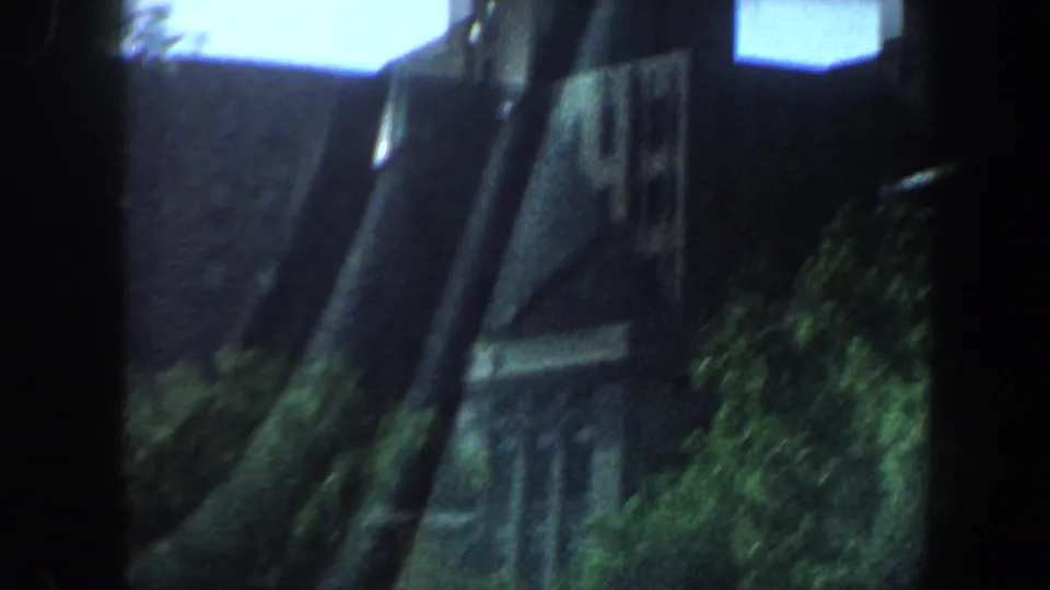 1964 Saint Louis frame showing an angled roofline or wall edge, trees, and shrubs as the camera continues past the building.