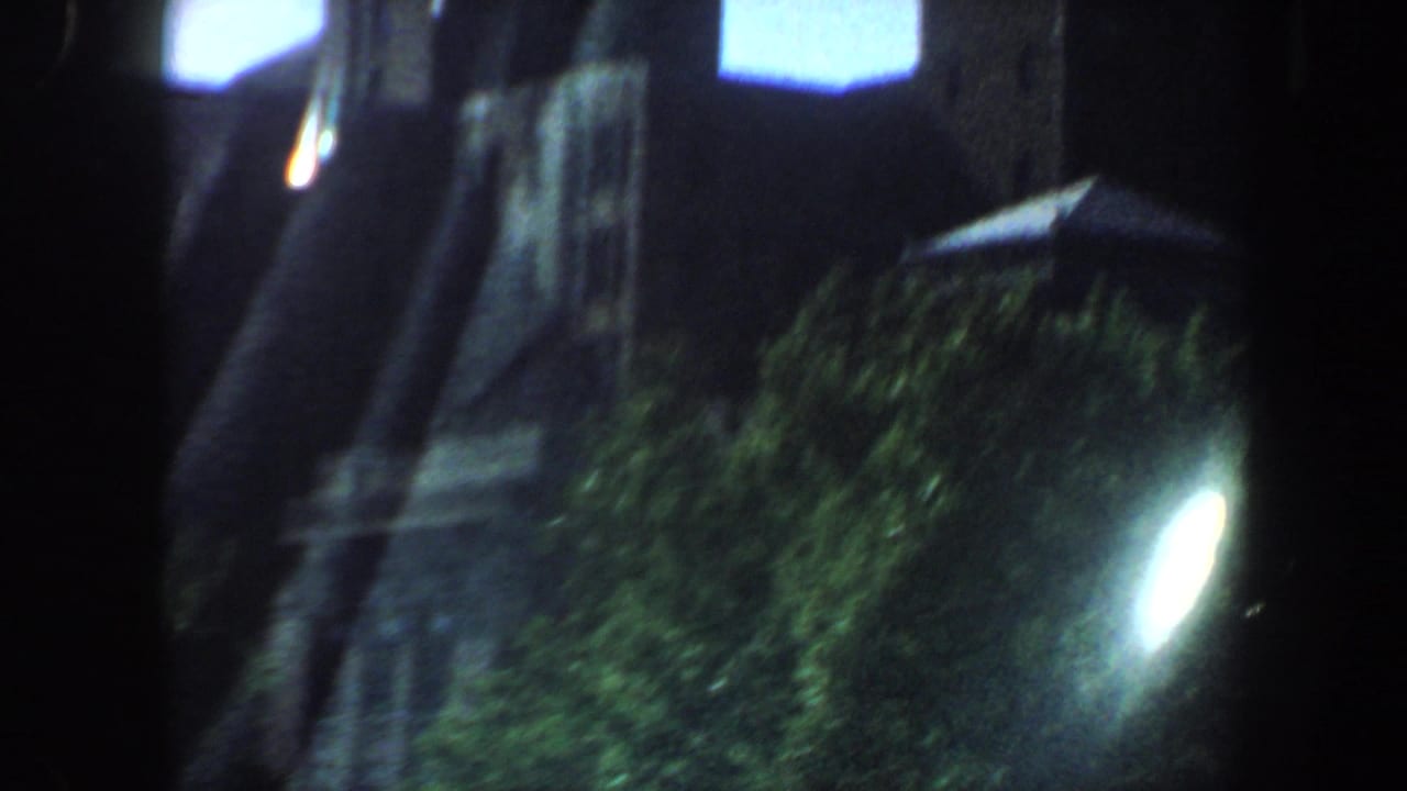 1964 Saint Louis home-movie frame showing a dim urban street edge with trees, dark buildings, and a bright light bloom.