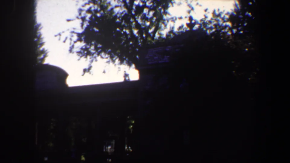 1964 Saint Louis home-movie frame with a house mostly in shadow behind tree trunks and leafy branches.